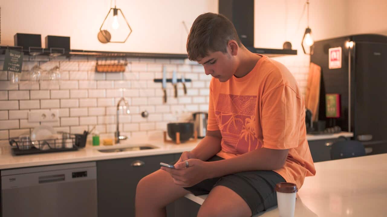 Teenage boy sitting at the kitchen table interacting with his smart phone while having a takeaway coffee