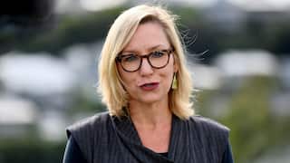 A blonde woman with black glasses and red lipstick speaking to a camera