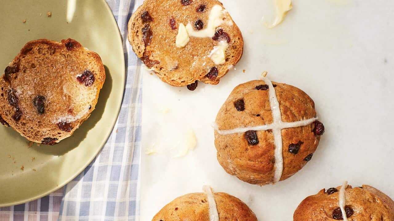 Seen from overhead, four hot cross buns, studded with fruit, sit on a white board, which is sitting on a blue-checked cloth. A fifth bun has been split and buttered, with one half sitting on the board and the other on a green plate that sits beside the board.