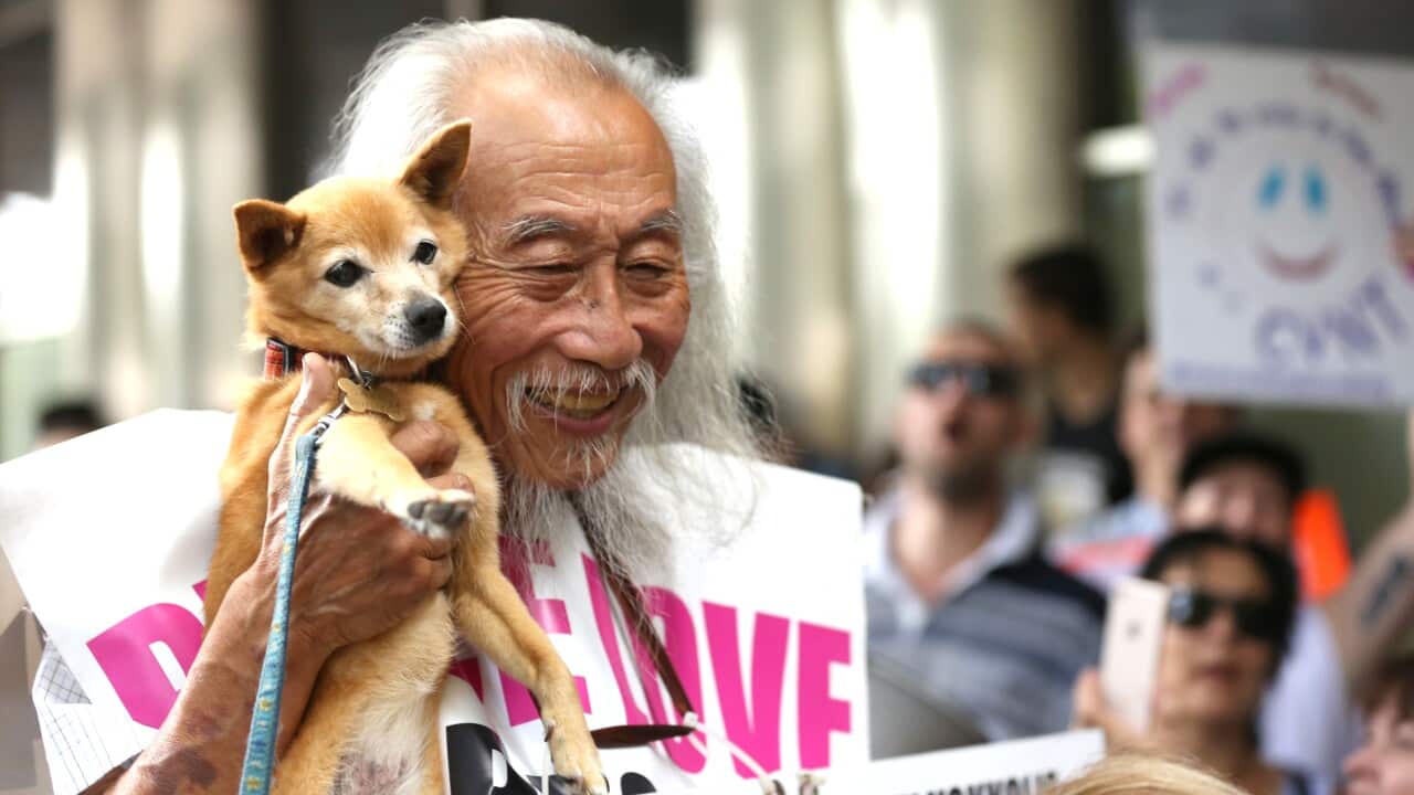 An old man smiling while wearing a sandwich board sign and holding a dog to his face.