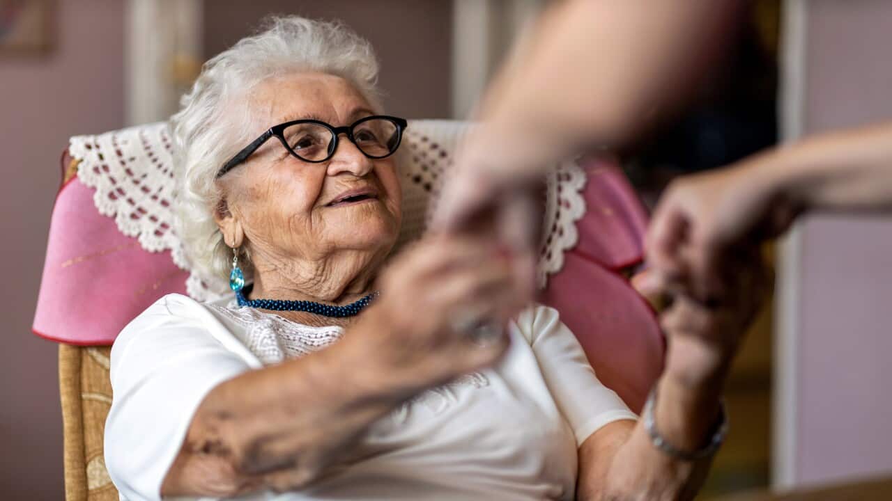 Female home carer supporting old woman to stand up from the armchair at care home