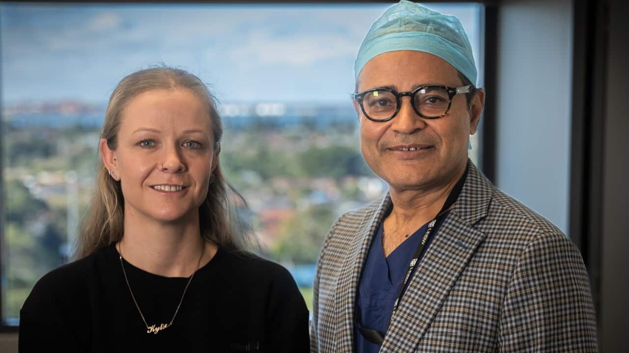 A woman in a black sweater stands in front of a window next to a man in a checked jacket.