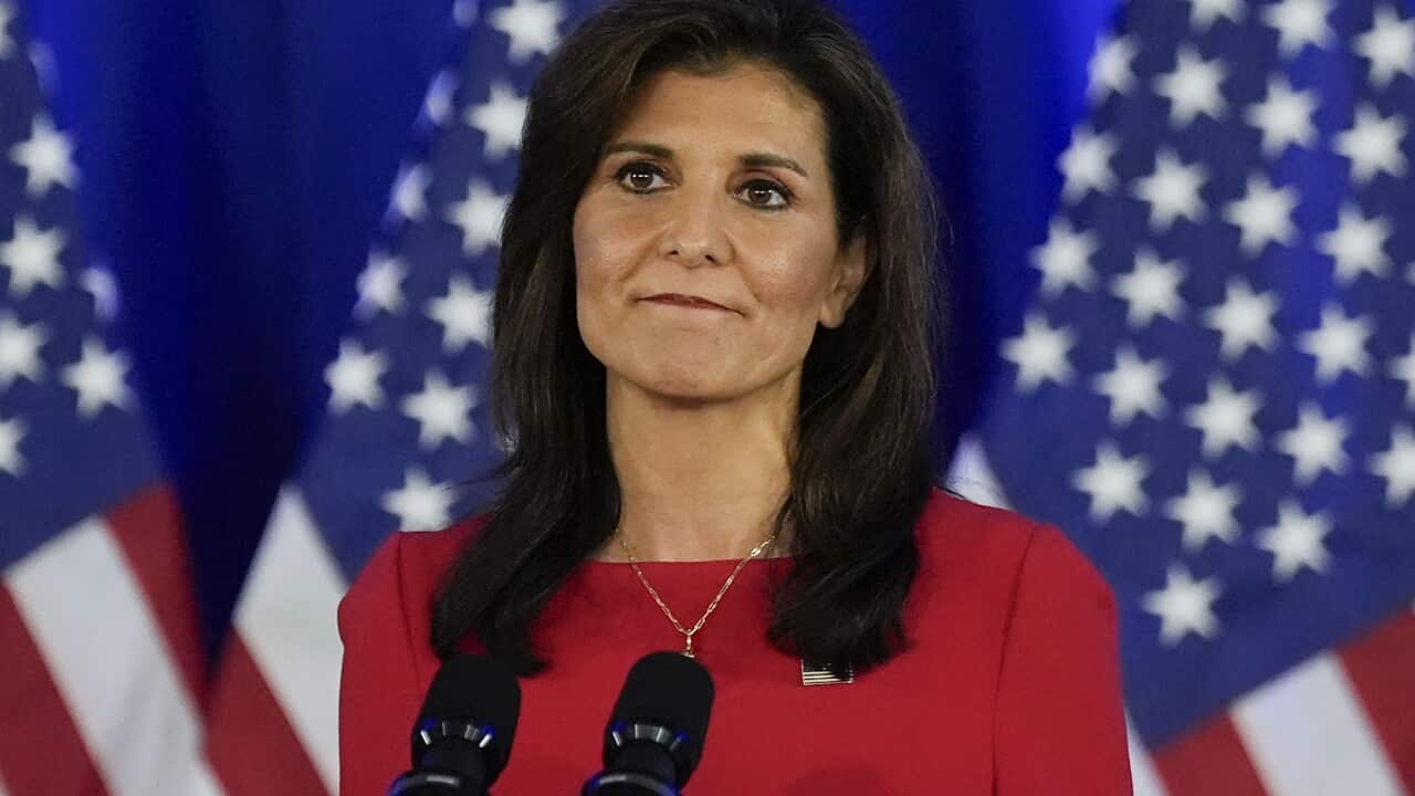 Woman with brown hair in red dress stands in front of microphones on a podium with red white and blue flags behind her.