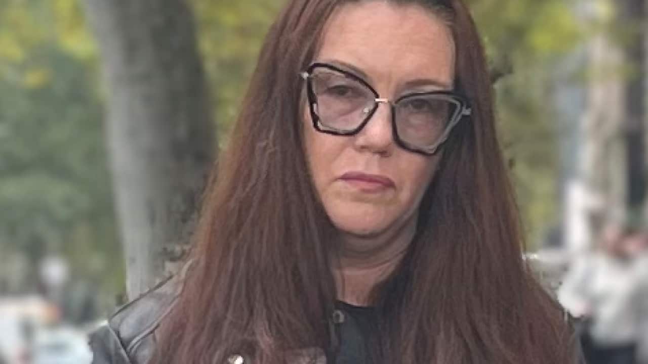 A woman wearing a white t-shirt, black jacket and glasses. 