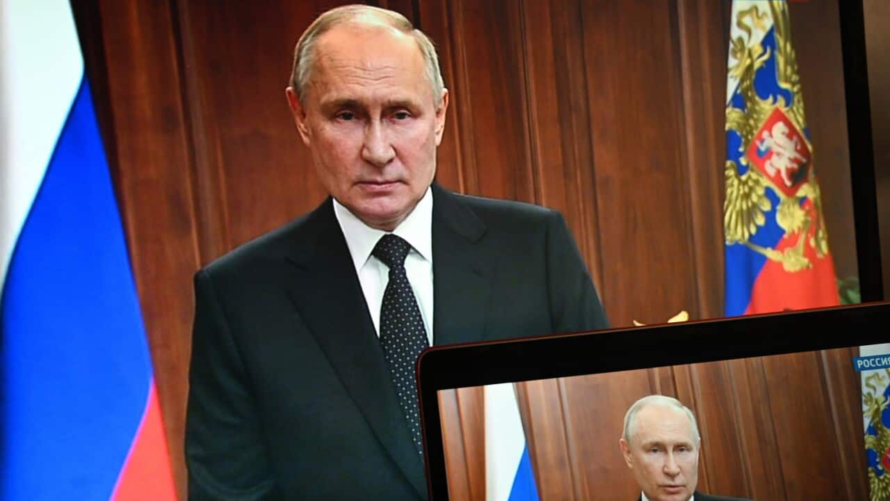 A man stands next to two flags. A monitor showing the same image is in the bottom left corner.