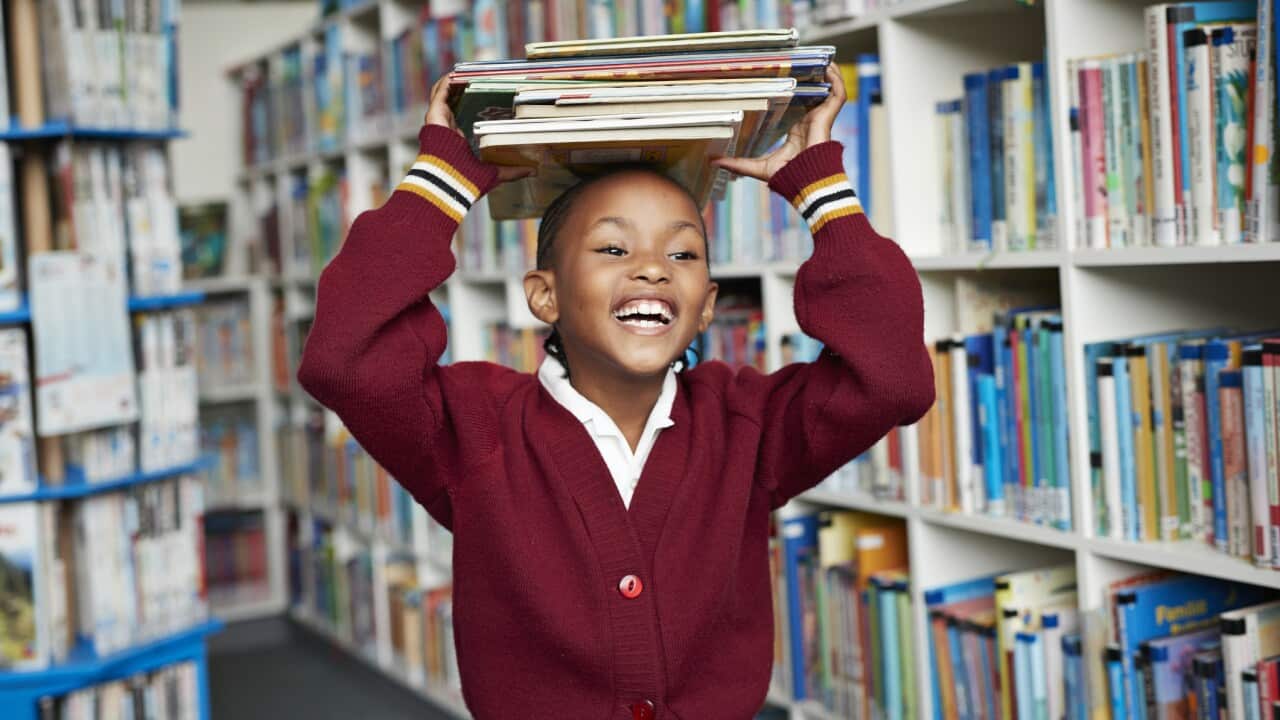 Cute schoolgirl smiling & balancing stack of books on the head at library