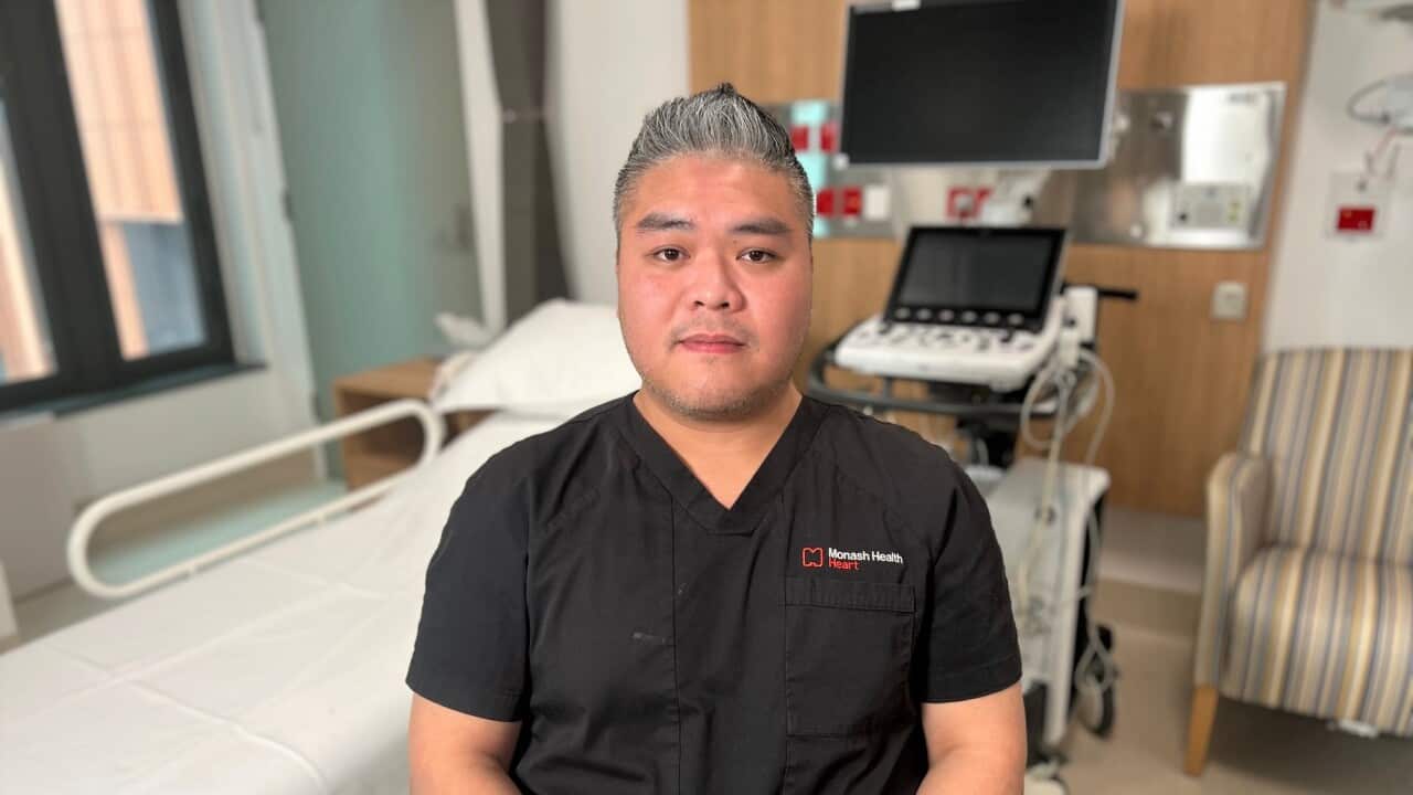 A man in a black nursing uniform sits in a hospital room, in front of medical equipment.