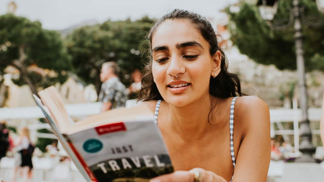 A woman lies on her stomach on a beach, and reads a travel guide