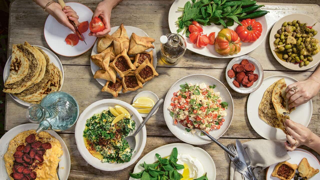 Seen from overhead, an array of Lebanese dishes sits on a wooden table. Hands can be seen folding a pastry at one end.