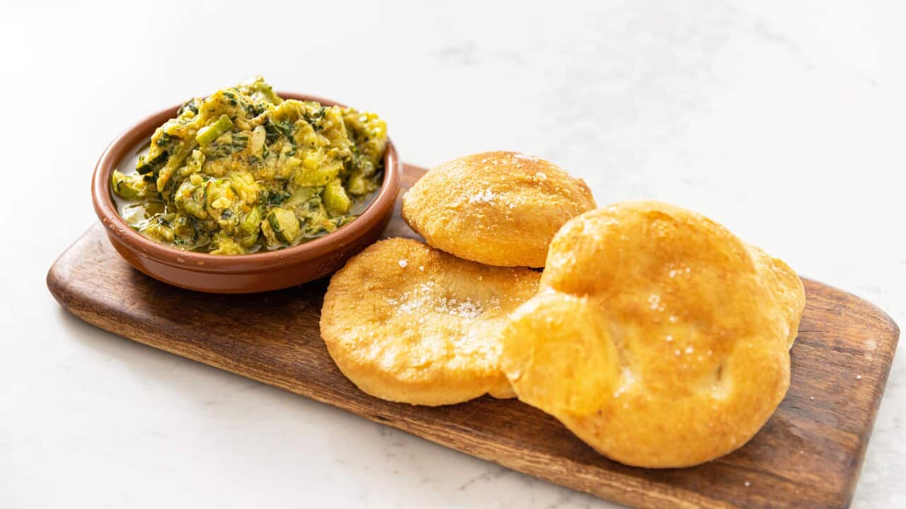 A brown pottery bowl full of green dip sits on long wooden board. Three golden roundish pieces of fried bread sit alongside the bowl.