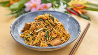 A beautiful blue-grey bowl sits on a wooden surface. It holds a pile of cooked noodles with egg, tofu and greens. A set of chopsticks sits to one side of the bowl. Fresh flowers can be seen in the background.