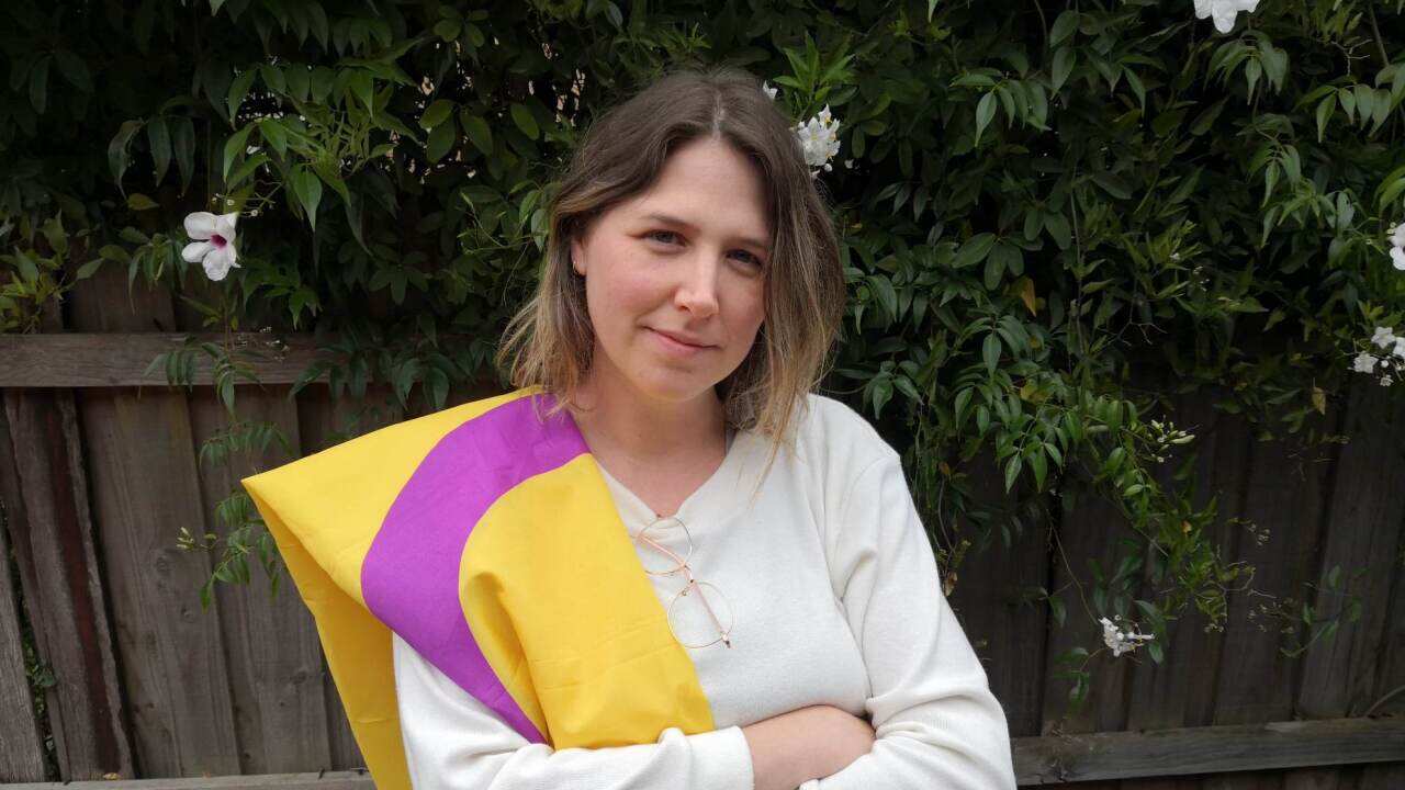 A woman stands in front of a wooden fence with a purple and yellow flag over her shoulder