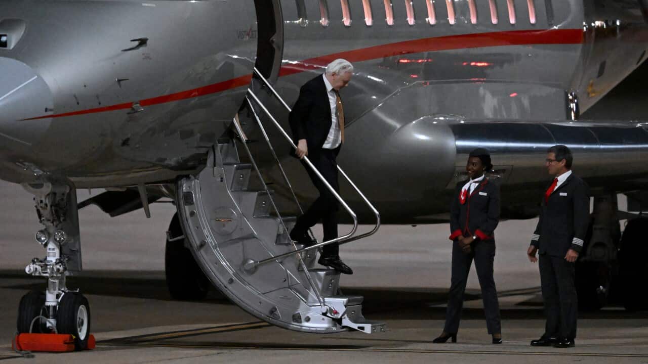 A middle-aged man with grey hair wearing a suit (Julian Assange) descends from a staircase out of a plane.