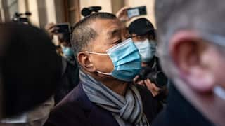 A close-up shot shows Jimmy Lai wearing a light blue surgical mask and a striped scarf as he walks through a crowded area surrounded by people with cameras and smartphones.