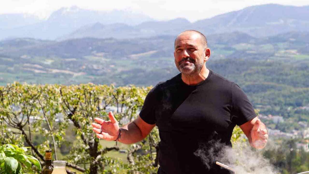 A man in a black t-shirt stands in a vineyard, with a saucepan on a portable stove on the table in front of him. Rolling mountain scenery can be seen behind him.