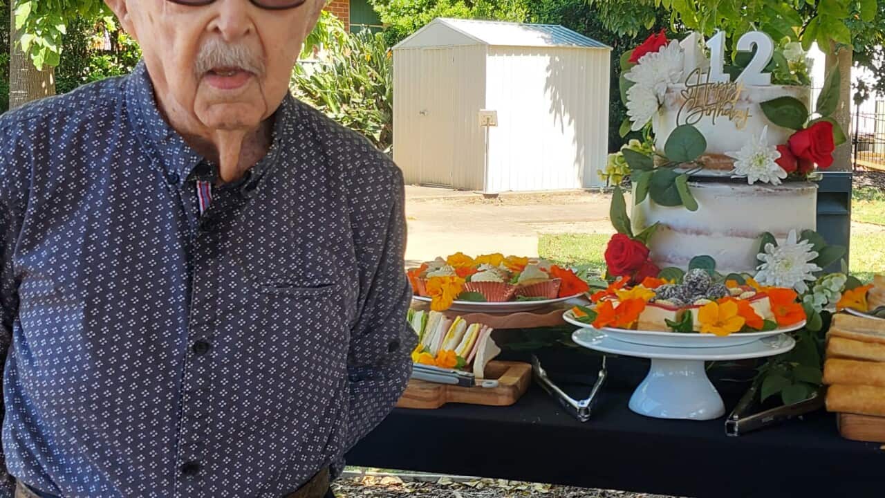 An elderly man stands in front of a table with a birthday cake that has “112“ candles on top.