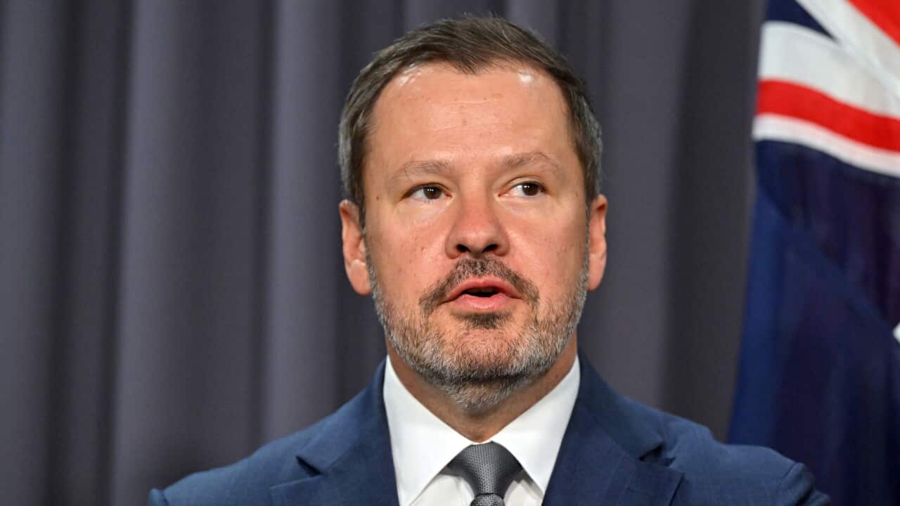 A man in a suit is standing in front of a blue curtain and an Australian flag.