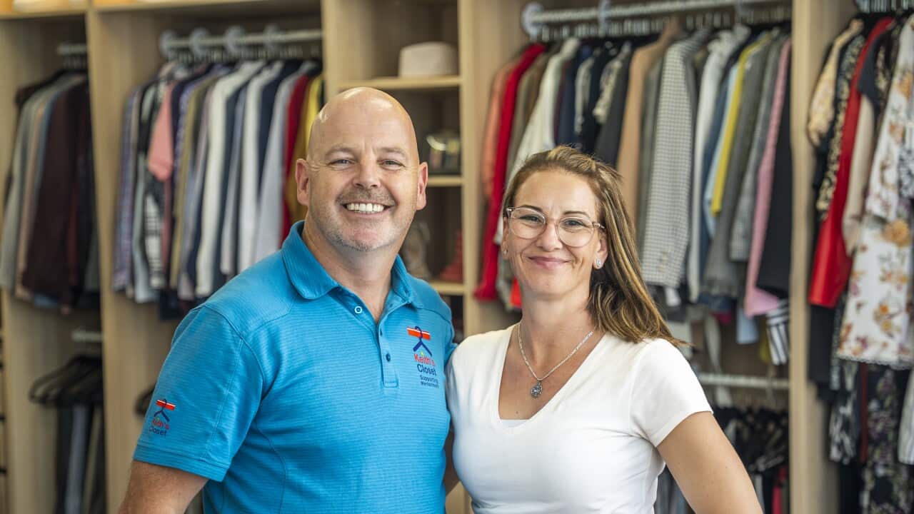 A man and a woman stand in front of a clothing display, smiling at the camera.
