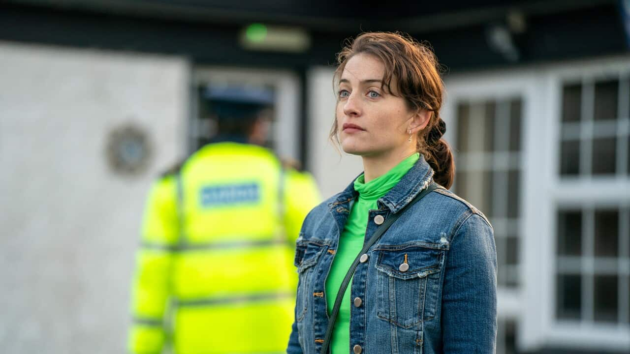 A pensive woman in a green top and denim jacket stands in front of a white building. A figure in a bright yellow police jacket can be seen behind her.  