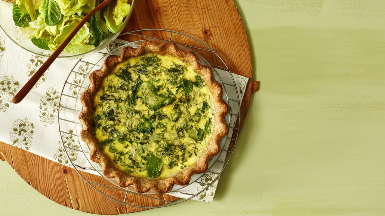 A large round woooden board sits on a green surface. On the board sits a bowl with leafy greens and beside it, a tart with fluted pastry and herby egg filling.