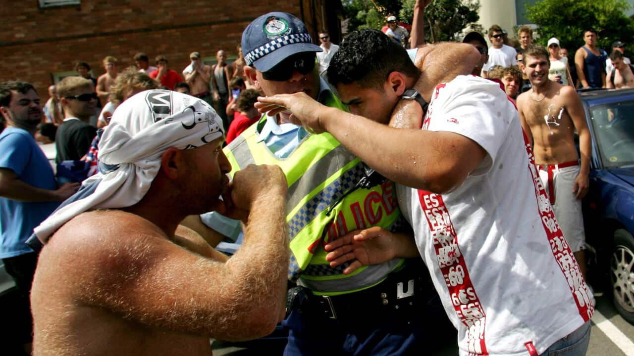 A police officer in a high-visibility vest intervenes between a shirtless man and a man in a white T-shirt fighting in a crowded, outdoor setting.