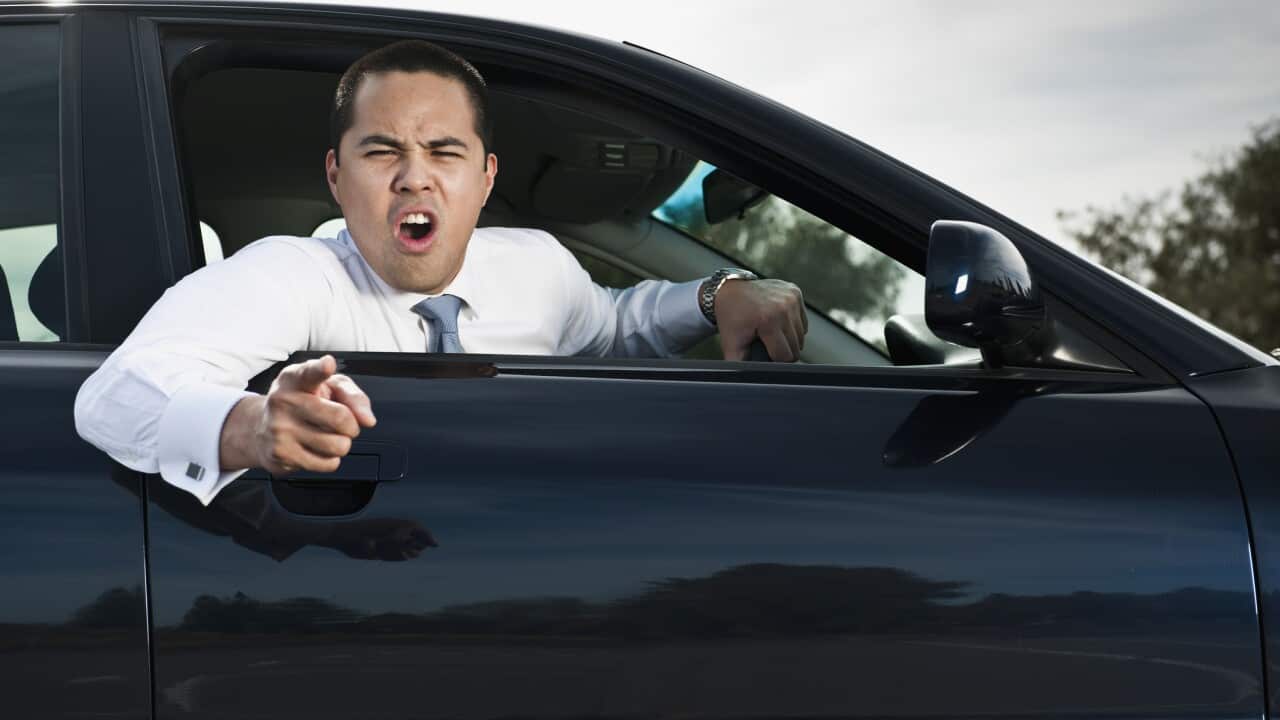 A man in a white shirt and silver tie leans out his car window and points and shouts