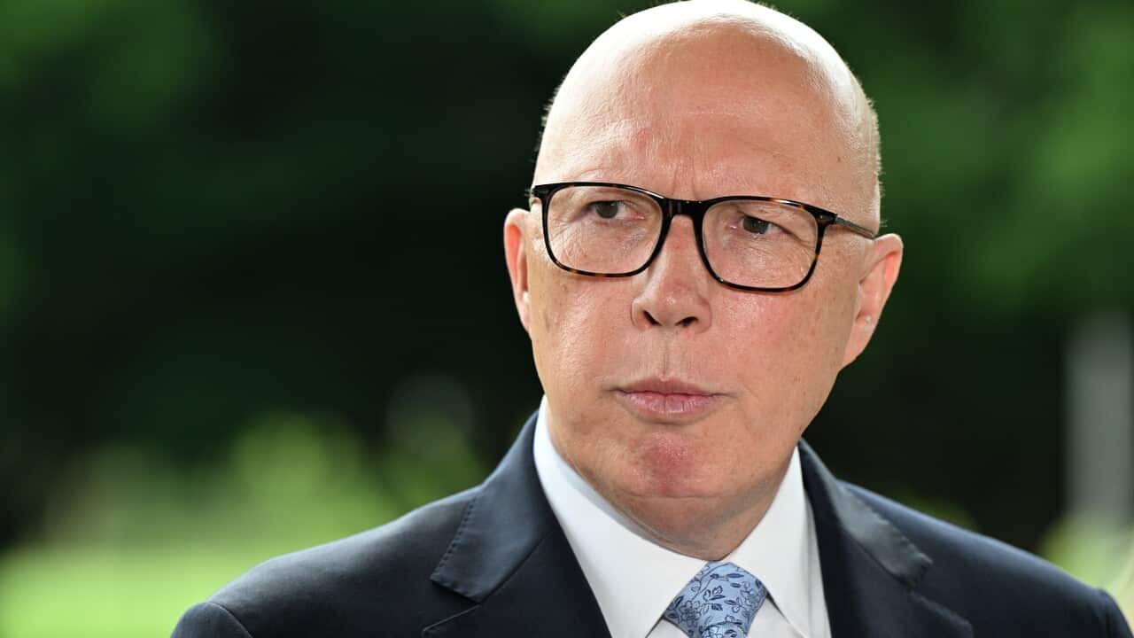 Opposition leader Peter Dutton wearing a suit, tie, and glasses is standing outside.