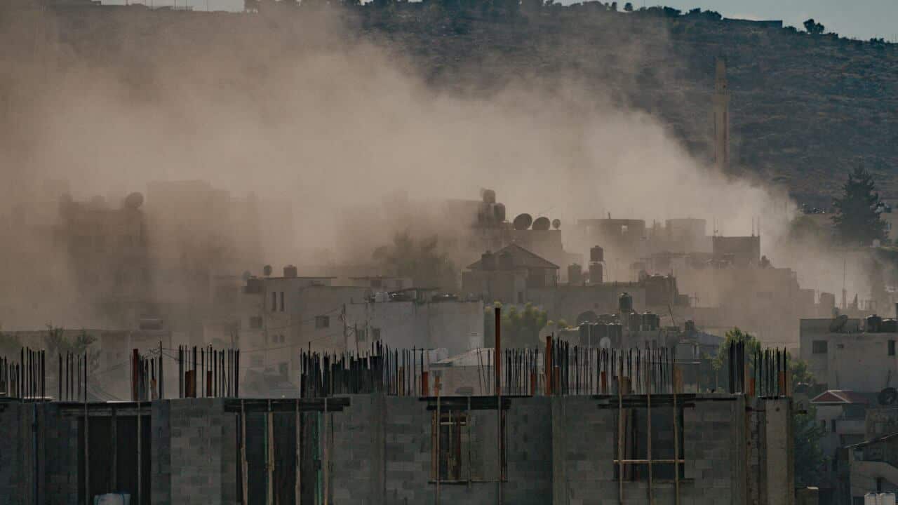 Smoke rises between densely packed buildings with a hilly landscape behind.