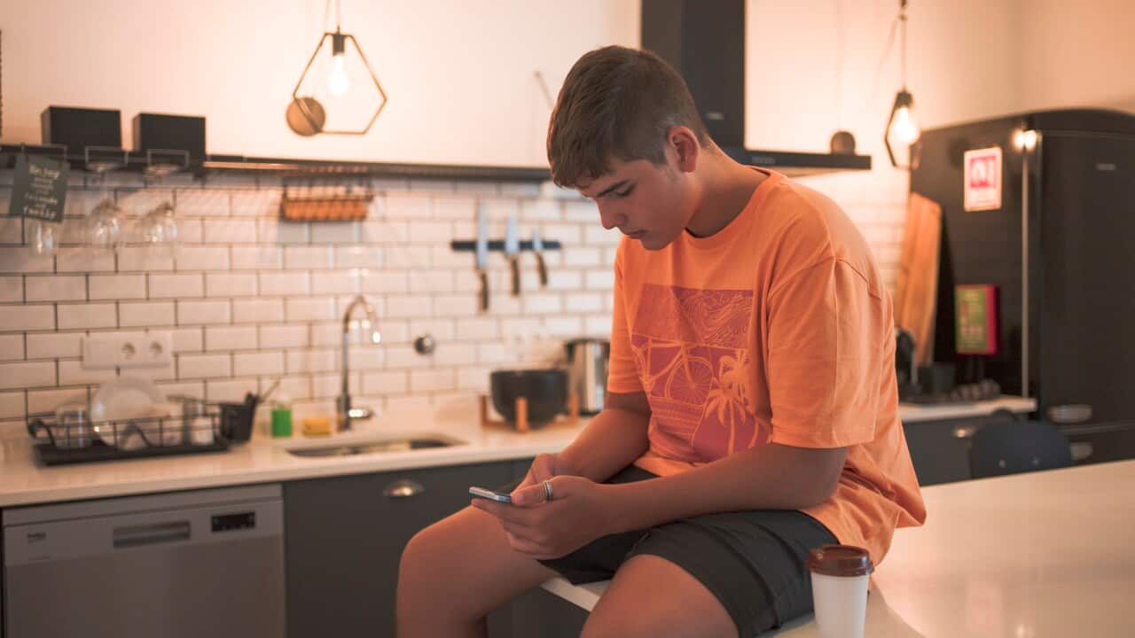 Teenage boy sitting at the kitchen table interacting with his smart phone while having a takeaway coffee