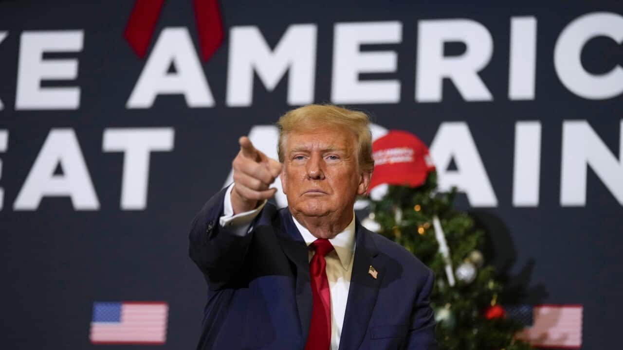 Donald Trump, wearing a blue suit and red tie, points at his supporters at a rally.