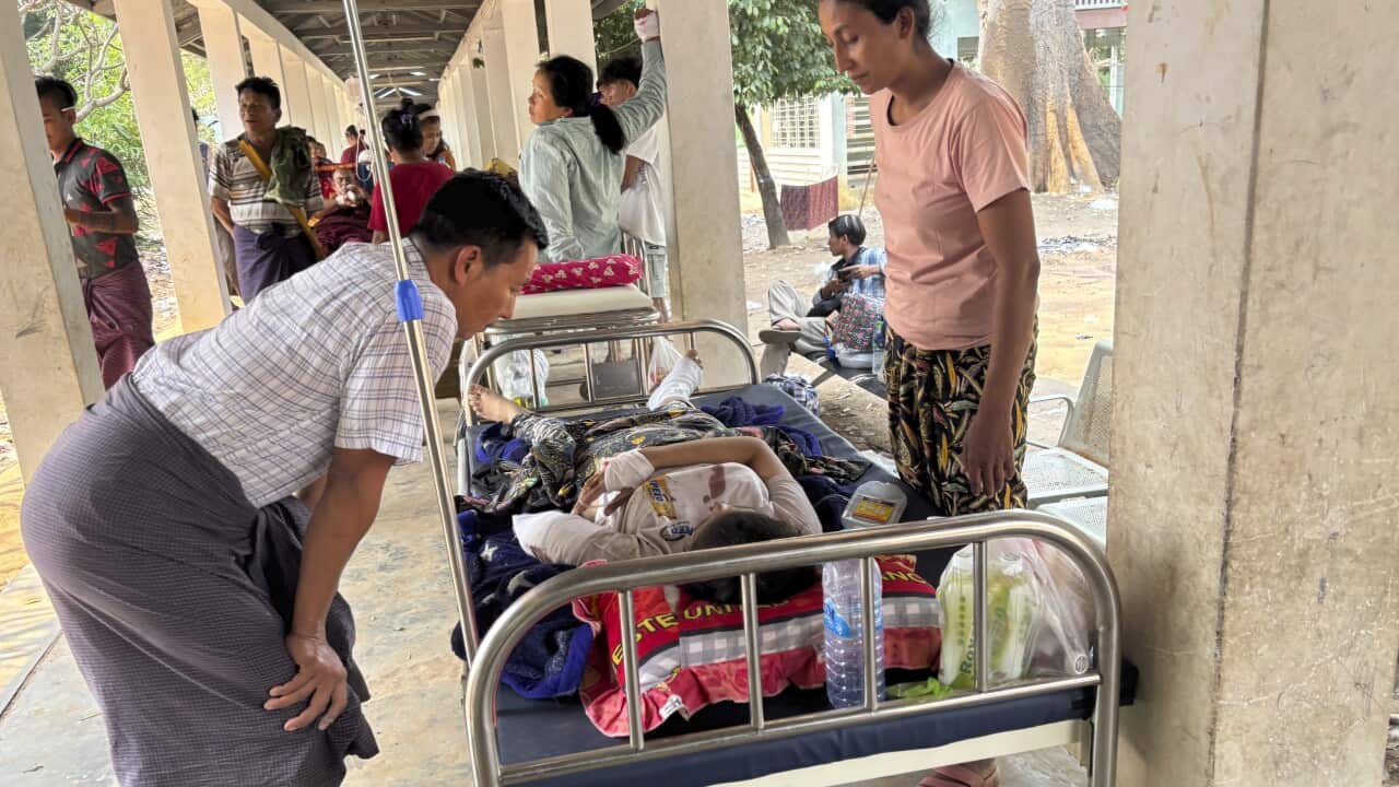 An injured person lying in a hospital bed, with two people standing over them. Others can be seen in the background.