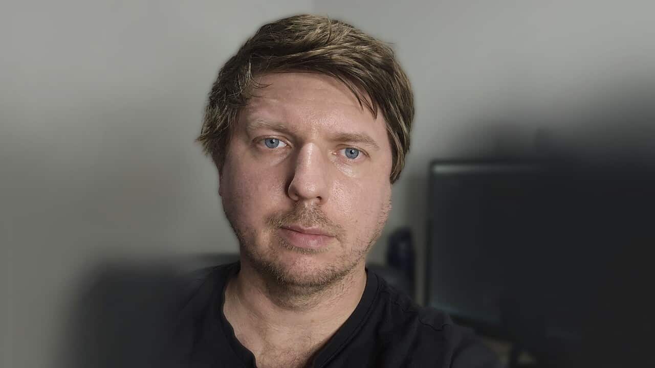 a man in his thirties takes a selfie at a desk that has a microphone and editing software open on his desktop
