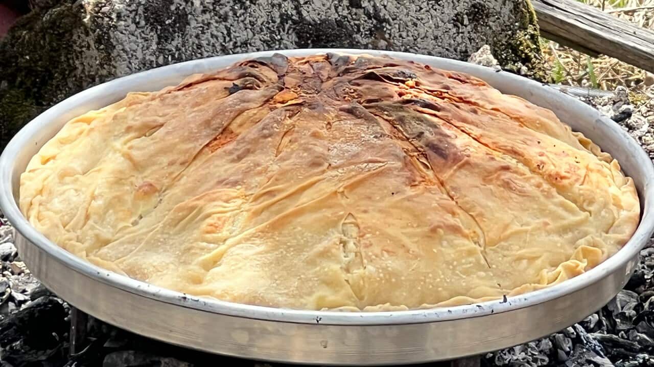 A large round silver pie tin with a cooked Greek pie in it sits above a pile of grey-black embers and ashes.