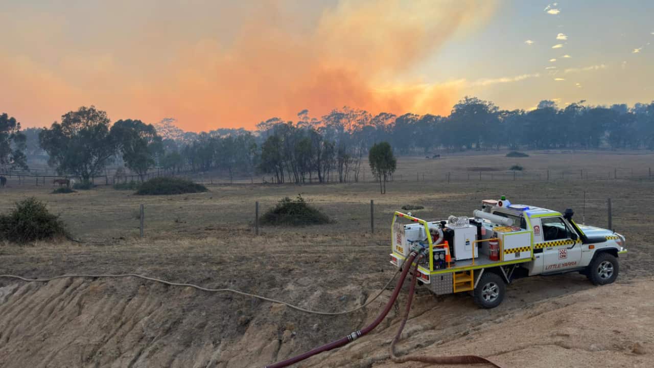 Smoke rises from the forest. A small firefighting vehicle is plugged into water pipes in the foreground.