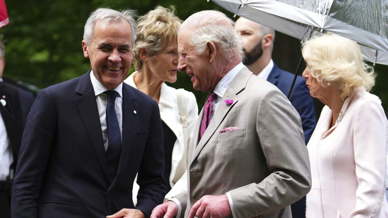 On the left, a man in a black suit smiles as he stands next to King Charles, with Queen Camilla behind them.