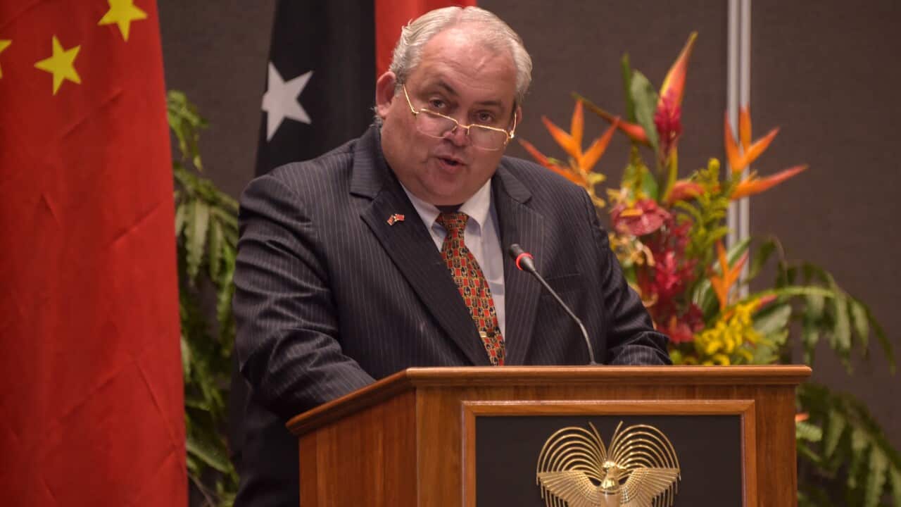 A man wearing a suit and wire-frame glasses speaks into a lectern microphone.