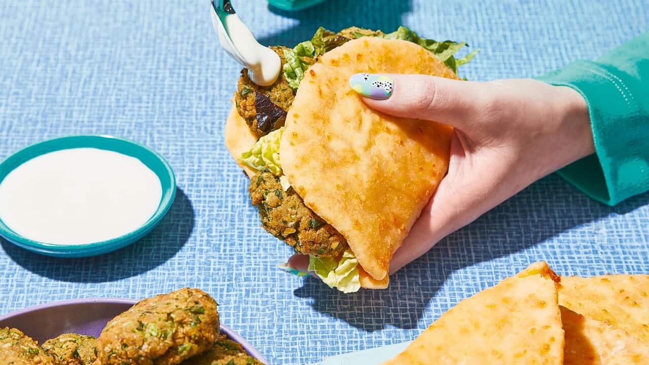 A hand holds a flatbread folded around a filling. A bowl of falafel and a board with more flatbreads sits on the table.
