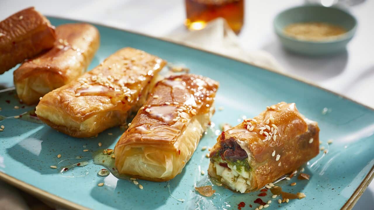 Four golden-brown filo parcels sit on a blue plate. One has been broken open to show the feta and pesto filling The parcels have been drizzled with a light golden honey sauce.