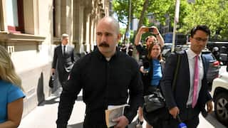 A white bald man with a moustache and wearing a black jumper walks down a city street. Reporters holding microphones and phones follow him.