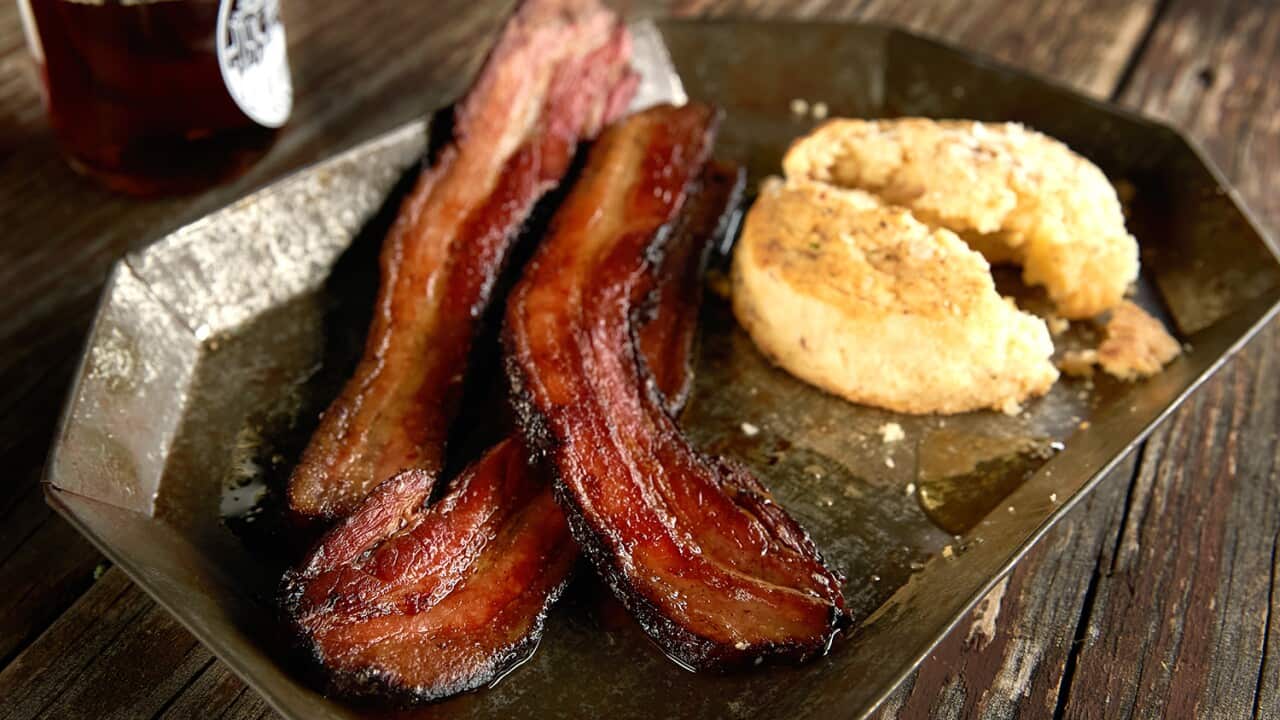 Three thick slices of cooked bacon site on a metal place, along with part of an American-style biscuit.