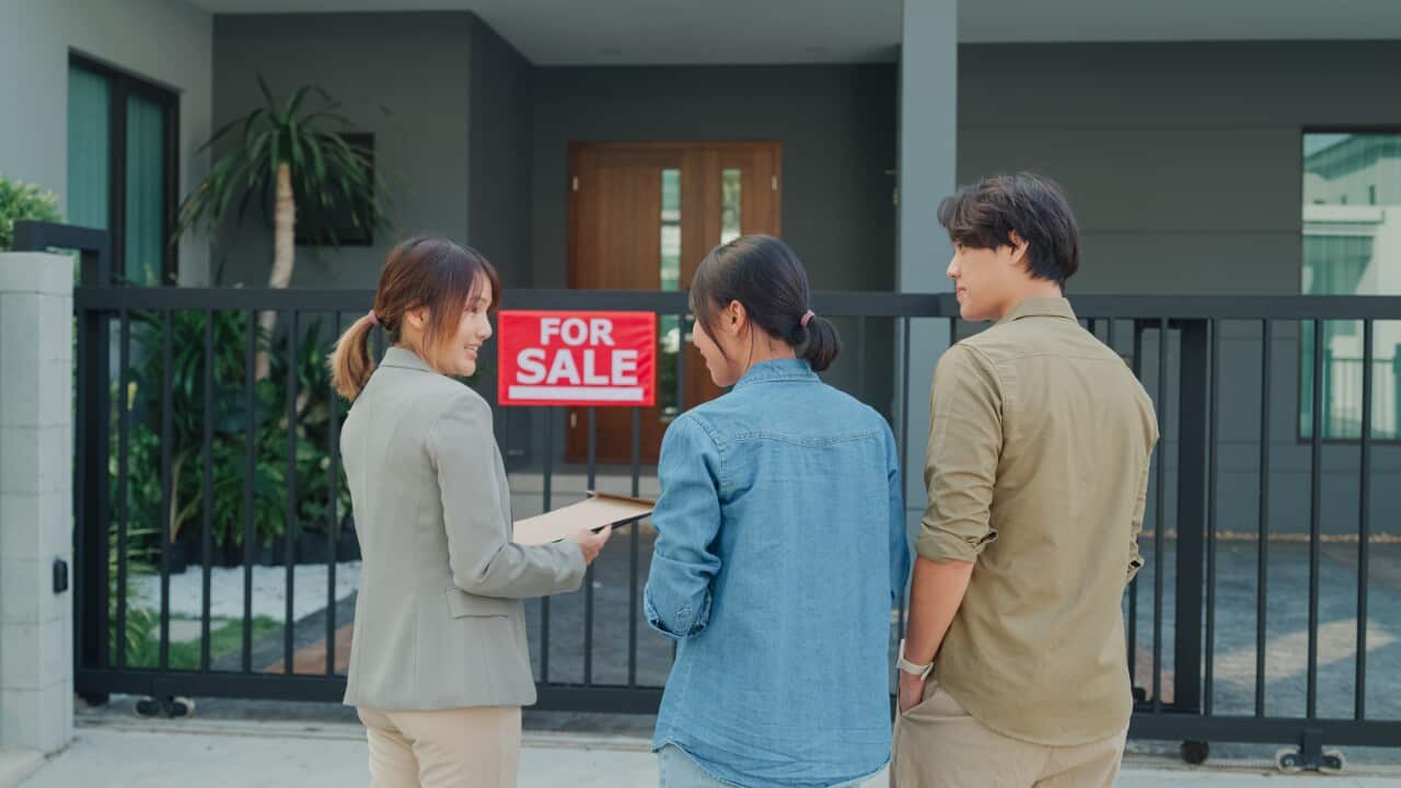 solo yachtswoman A young female real estate broker shows a house to potential buyers outside at house with a 'For Sale' sign on the gate.