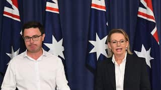 A woman speaks at a press conference with a man seen standing in the background.