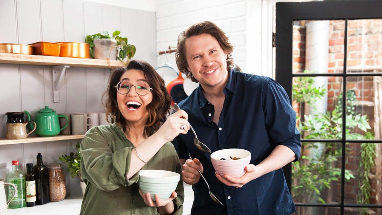 A man and a woman stand in a kitchen, with full-length glass door showing greenery behind them. They both hold bowls and spoons, and appear to be laughing happily.