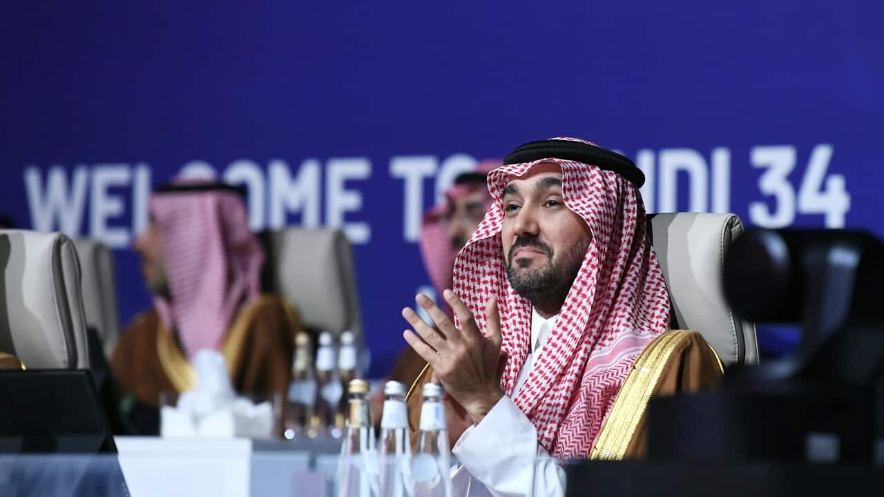 A man wearing a Saudi-style keffiyeh looks forward with a smile while rubbing his hands together.