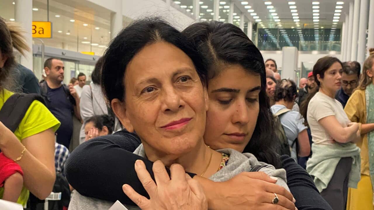 A young girl hugs an older woman from behind. The older woman is wearing a grey sweater.