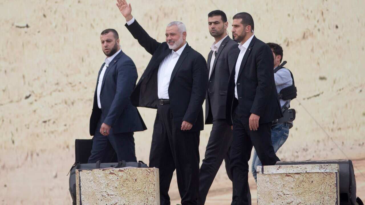 A group of men wearing suits walk together. One middle-aged man with a white beard raises his right hand.