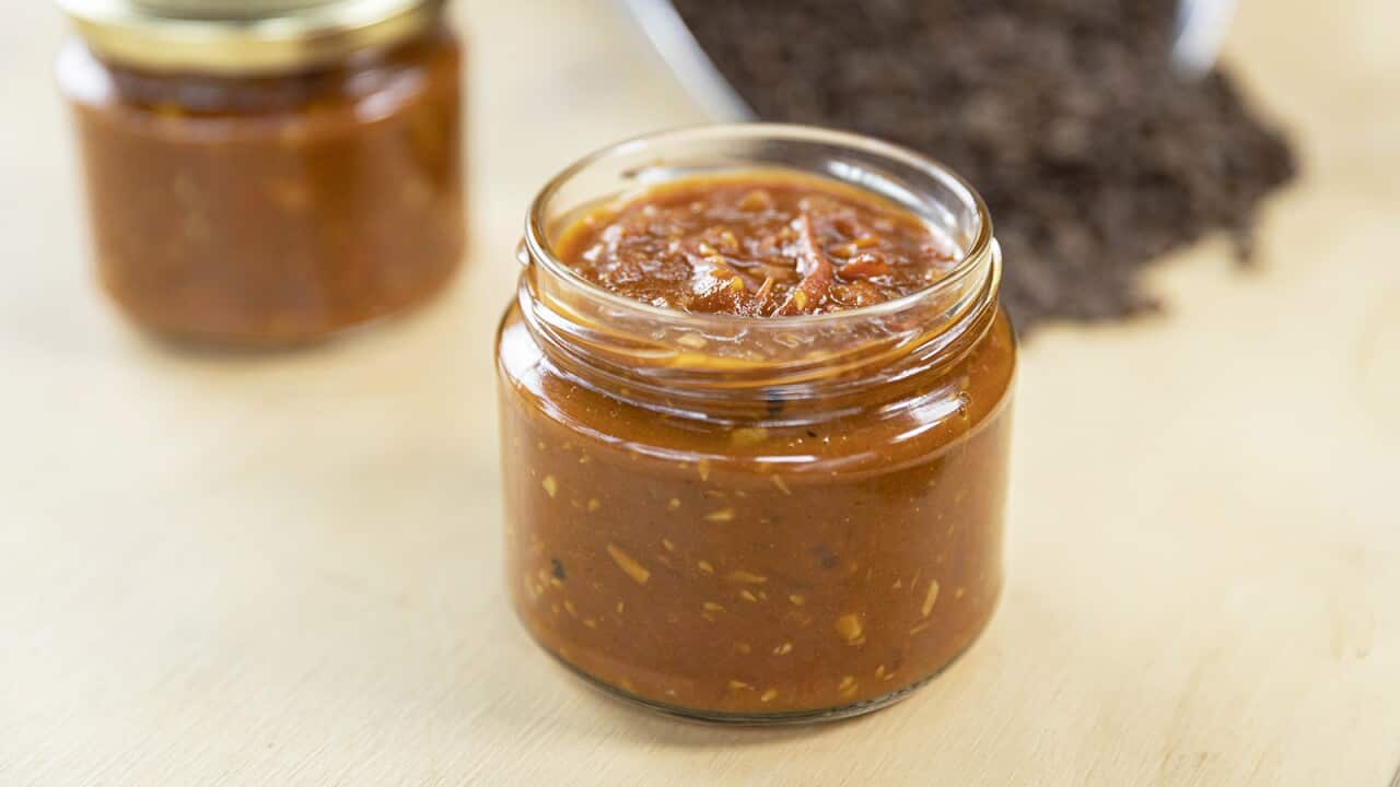 A glass jar holds a reddish brown sauce. Another jar of sauce, this one with a gold lid, sits behind it. A scoop holding coffee beans can be seen in the background.