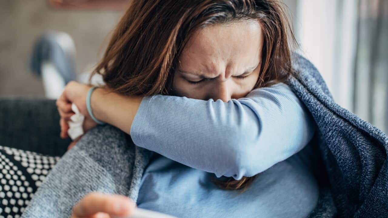 Woman sneezing into her elbow sitting on sofa and holding thermometer
