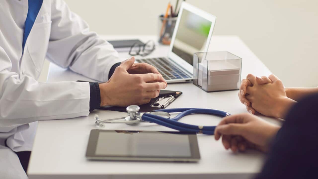 A doctor and two patients are sitting at a table during a consultation. Only their hands are visible. The doctor's hands are on a table beside a stethoscope, tablet and laptop. He is wearing a white doctor's gown. The patients' hands are visible in front of the doctor.