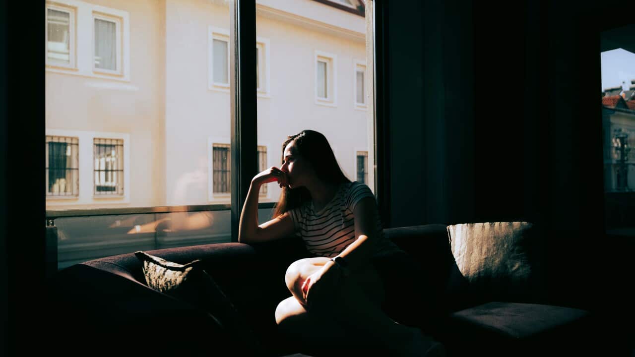 A woman sits in darkness on a sofa looking out a window